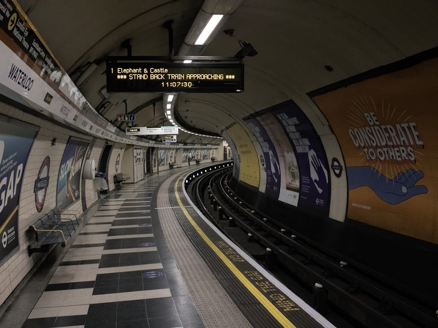 Inside Elephant and Castle underground station, the curved platform features black and white checkered floor tiles, with a row of empty metal benches along the tiled wall. Above, a digital display board indicates the approaching train to 'Elephant & Castle' with a timestamp of 11:07:30 and a message to 'STAND BACK TRAIN APPROACHING'. The station's tunnel walls are adorned with various posters, including one with the message 'BE CONSIDERATE TO OTHERS' depicted in bold letters and a graphic of a hand and a heart. Track rails run along the edge of the platform, curving into the tunnel, with safety line markings in yellow and white visible on the platform edge. The lighting is provided by ceiling-mounted fluorescent fixtures, casting a neutral tone over the scene. The station is quiet, with no passengers present, and the overall environment reflects a typical moment during the off-peak hours of the underground service, as part of the infrastructure supporting London house removals and packing logistics from nearby service providers.