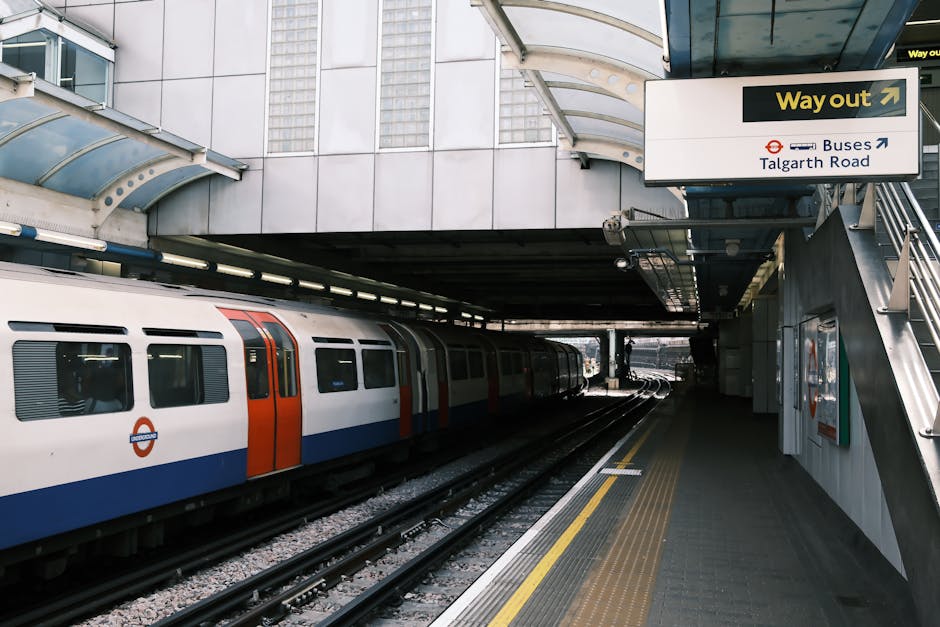 Inside Elephant and Castle underground station, the curved platform features black and white checkered floor tiles, with a row of empty metal benches along the tiled wall. Above, a digital display board indicates the approaching train to 'Elephant & Castle' with a timestamp of 11:07:30 and a message to 'STAND BACK TRAIN APPROACHING'. The station's tunnel walls are adorned with various posters, including one with the message 'BE CONSIDERATE TO OTHERS' depicted in bold letters and a graphic of a hand and a heart. Track rails run along the edge of the platform, curving into the tunnel, with safety line markings in yellow and white visible on the platform edge. The lighting is provided by ceiling-mounted fluorescent fixtures, casting a neutral tone over the scene. The station is quiet, with no passengers present, and the overall environment reflects a typical moment during the off-peak hours of the underground service, as part of the infrastructure supporting London house removals and packing logistics from nearby service providers.