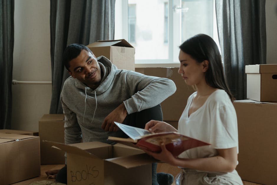 Inside a room during a home relocation, a man and a woman are seated on the floor among stacked cardboard moving boxes. The man, wearing a light grey hoodie, is leaning forward listening attentively, while the woman, dressed in a white top, is holding a booklet or folder and appears to be explaining or reviewing moving details. The boxes surrounding them vary in size and are some labelled, such as one marked 'BOOKS.' The room is illuminated by natural light coming through a window with dark curtains, and some boxes are open, possibly containing packed household items. Visible in the background are additional storage boxes, a cardboard box with an open top, and plastic packing materials. This scene captures a moment of preparation for a house move, highlighting the packing and planning process involved in a local flat move, with [COMPANY_NAME] providing moving services for such relocations, emphasizing the logistical aspects of furniture transport and packing.